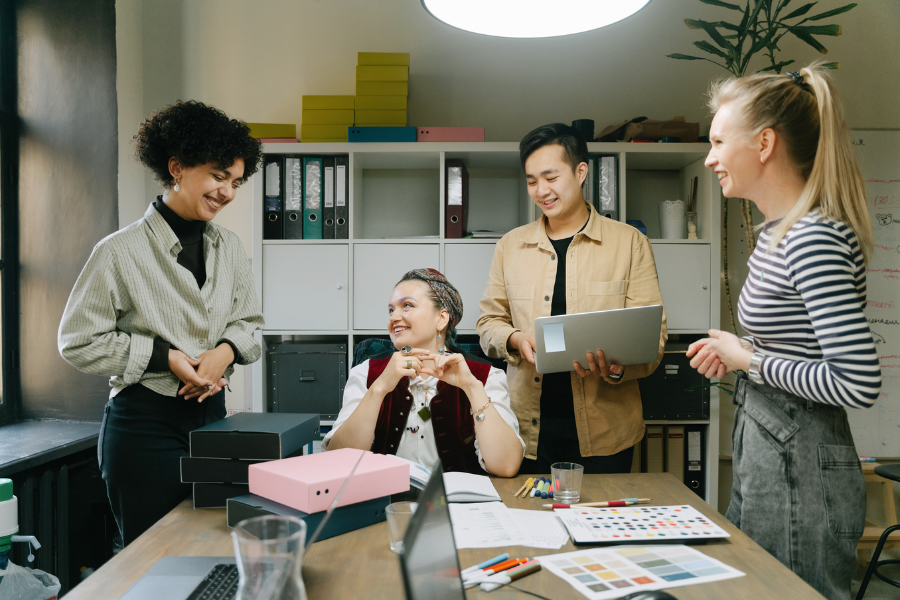 Four colleagues cheerfully discuss around a table in a bright office, with a laptop and color swatches nearby.